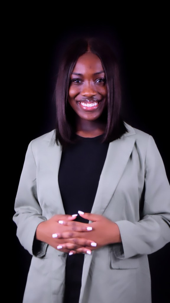 A woman with long dark hair smiles confidently while wearing a light green blazer over a black top, with her hands clasped in front of her.